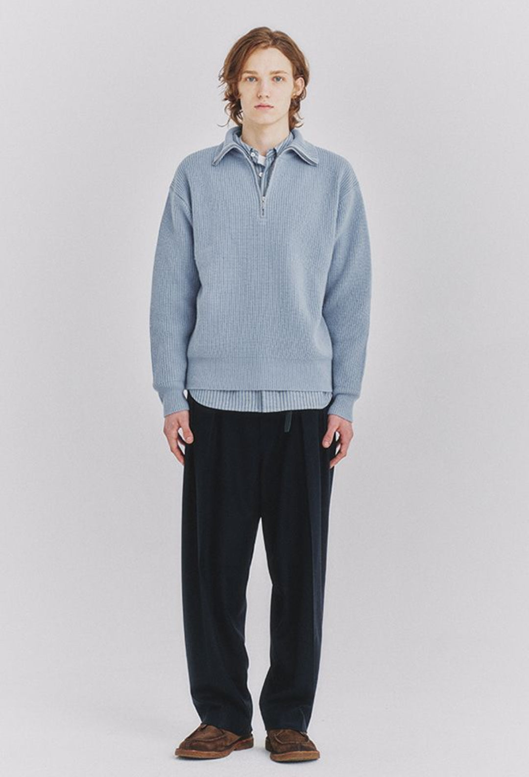 Person wearing a light blue sweater over a collared shirt with dark pants on a white background